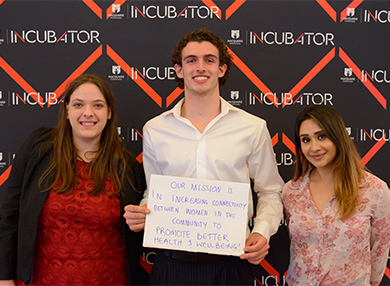 Student entrepreneurs hold a paper sign in front of the Incubator logo patterned wall