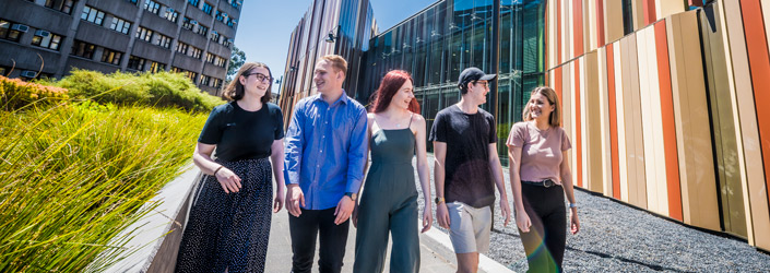 Five young adults smiling and speaking to each other while walking by the Macquarie Library.