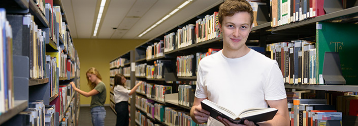 Male student reading in library with students choosing books behind him