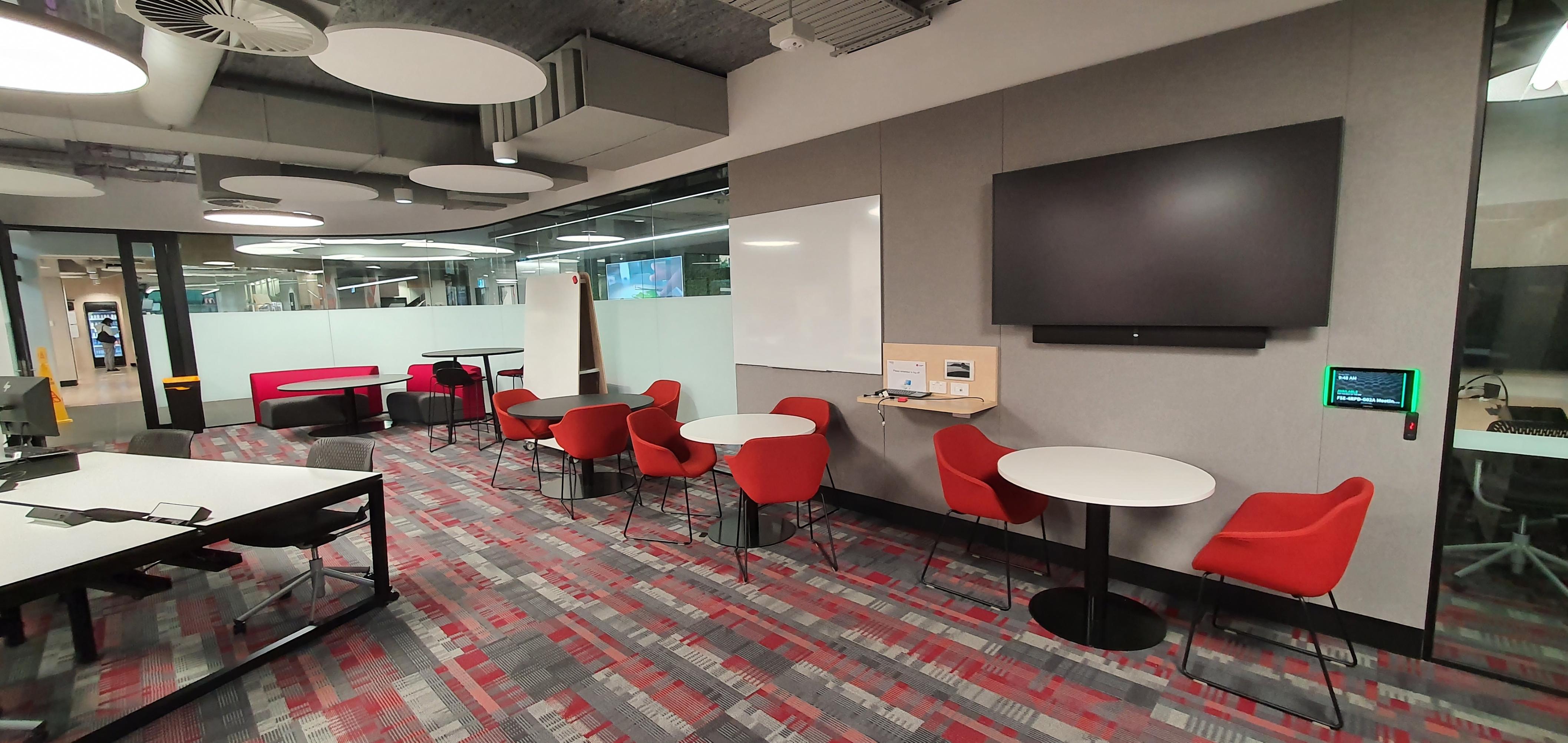 The drop-in centre at 4 Research Park Drive. It is a large, brightly-lit room with several small tables surrounded by chairs. There are white boards and screens mounted on the wall.