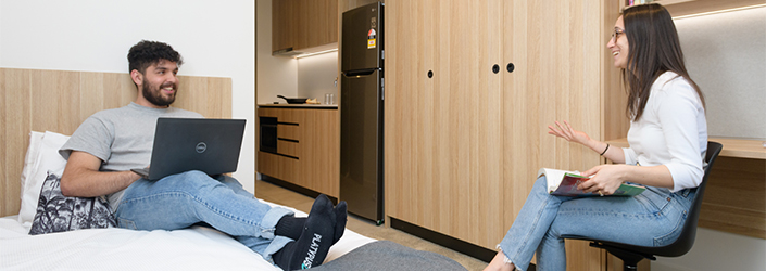 Student using a laptop while resting on a bed talking to another student sitting on a chair